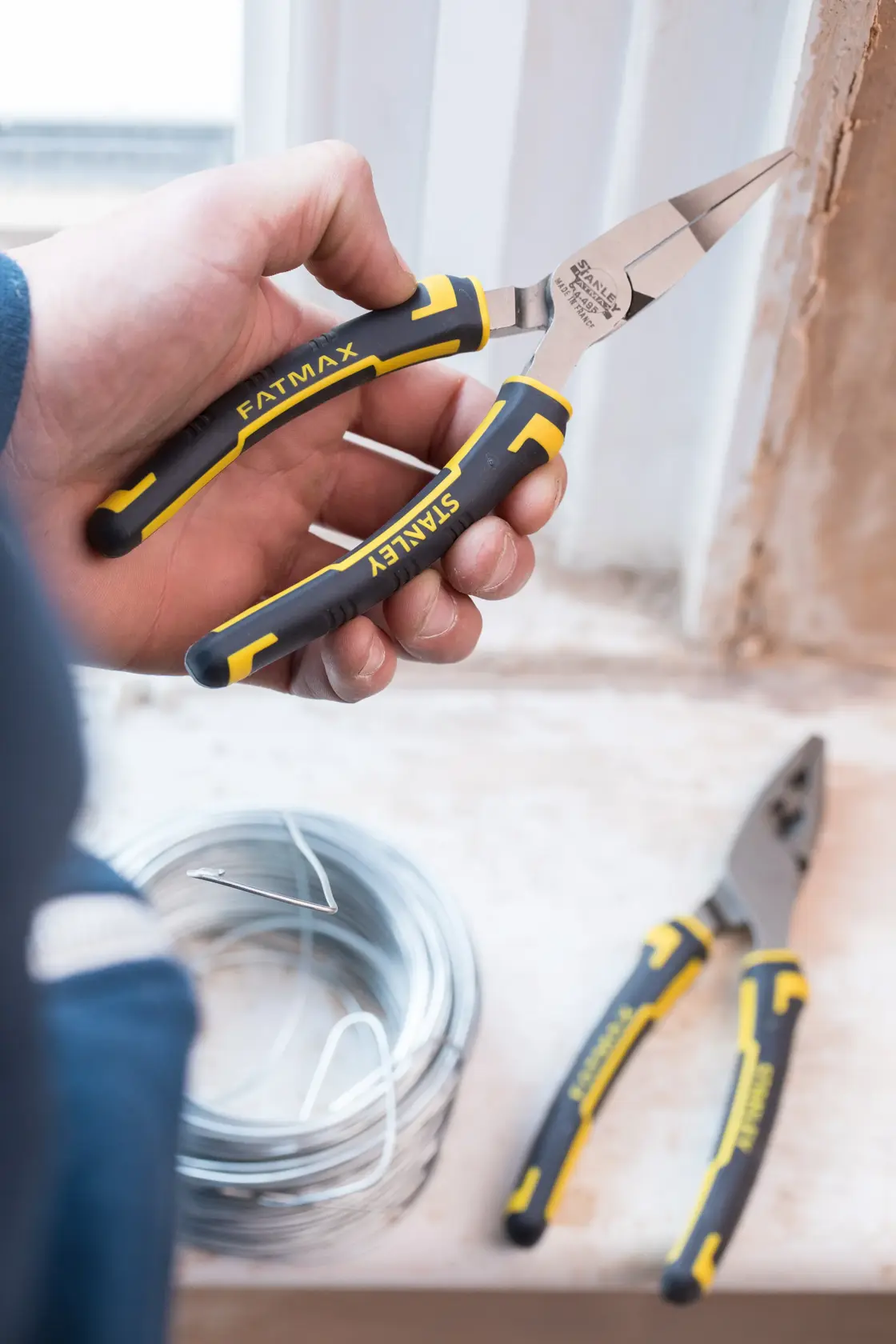 STANLEY 0-84-495 long nose pliers with yellow and black grips, held in hand near coiled wire, close-up.