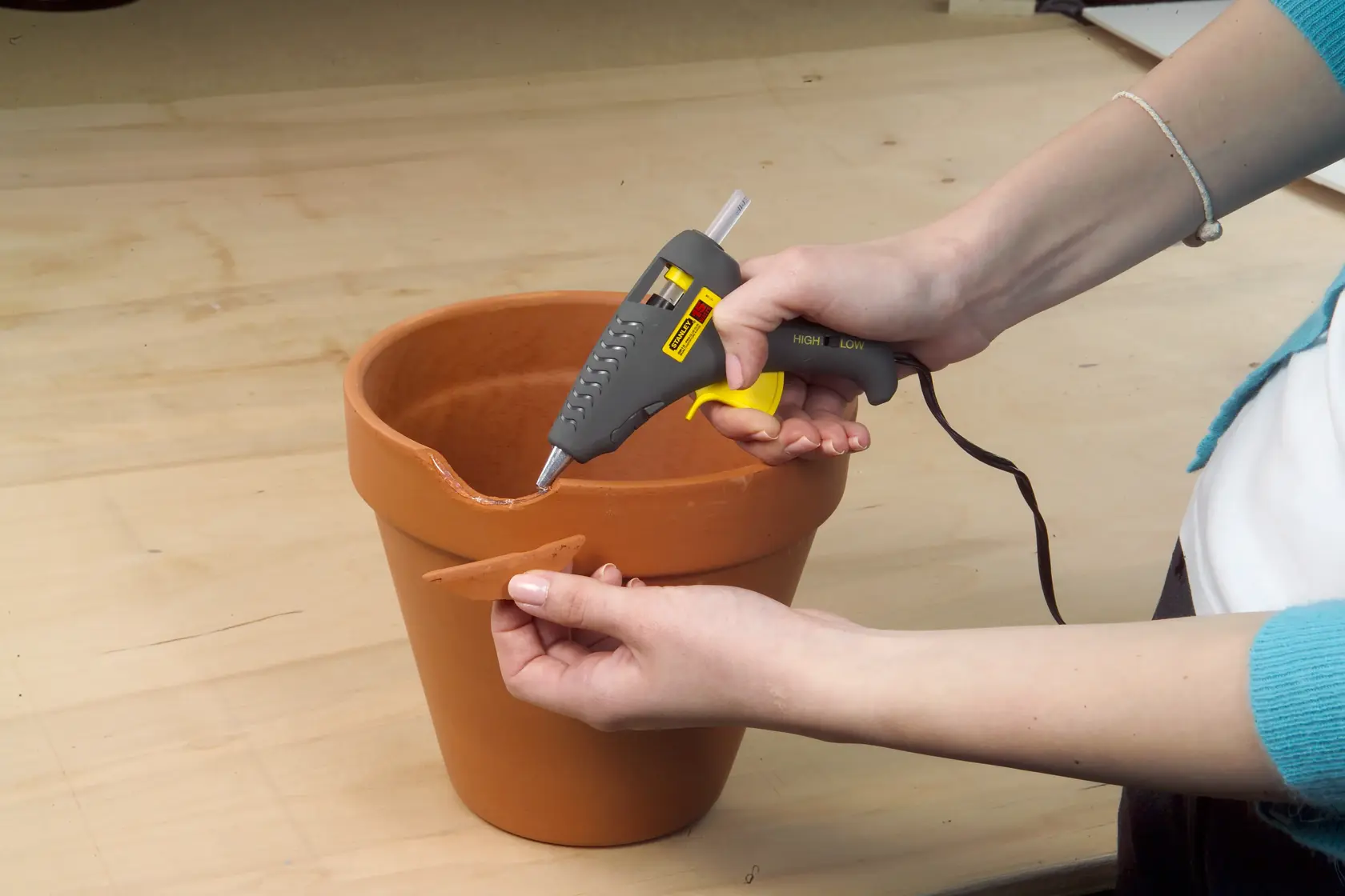 STANLEY 0-GR15 glue gun in hand applying hot glue to a broken terracotta pot on a wooden surface.