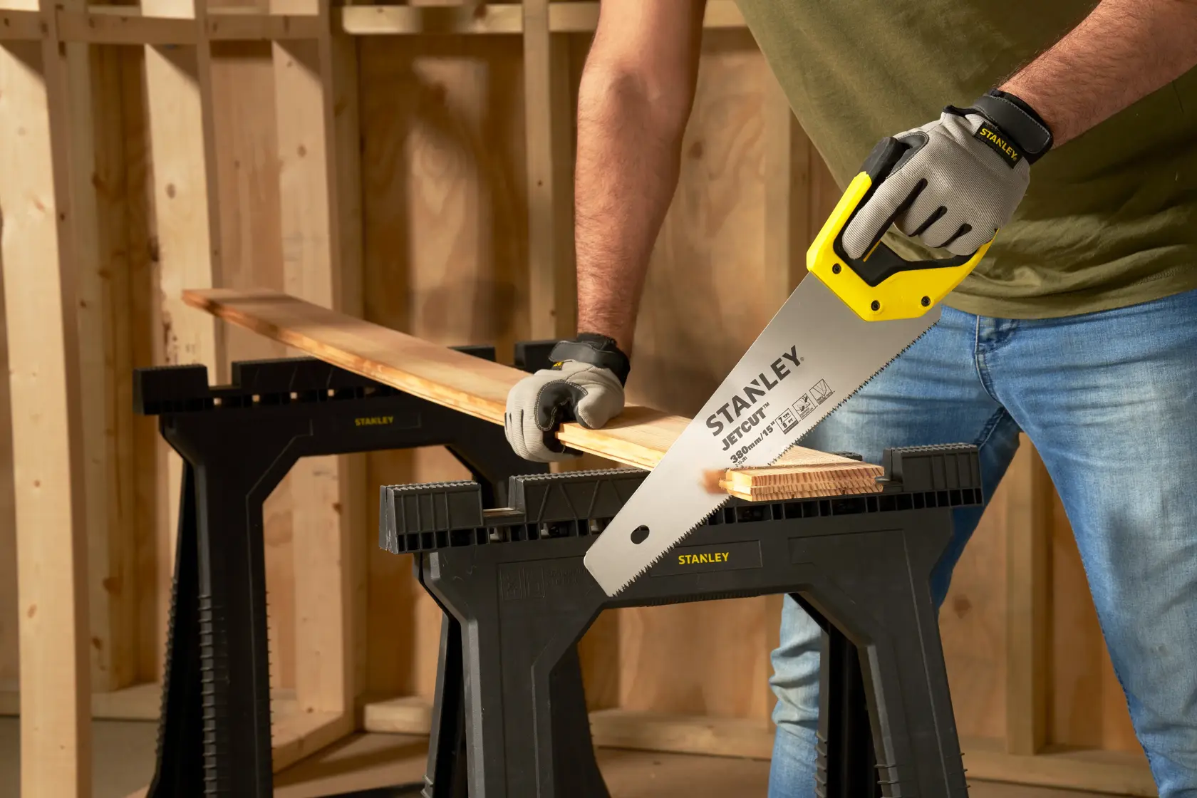 STANLEY 2-15-281 hand saw with yellow handle cutting wood plank on black saw horse in workshop.