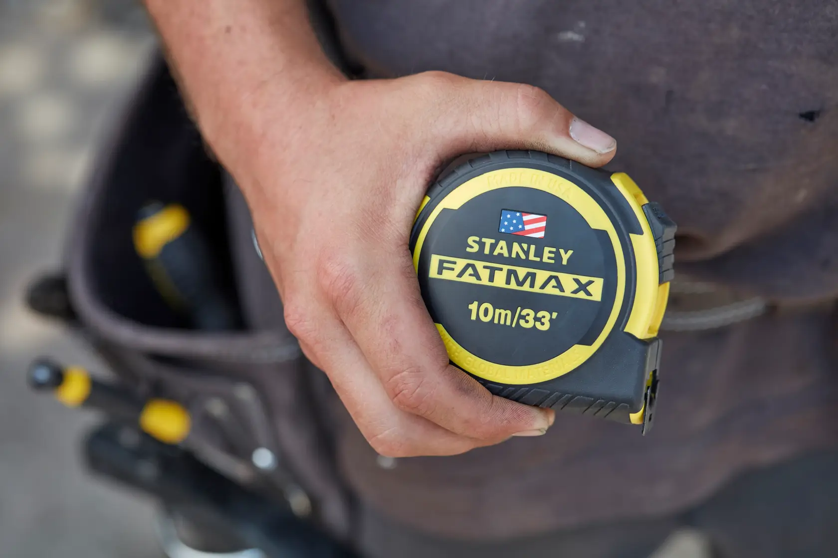 Photo of STANLEY FMHT0-36336 FATMAX 10m/33' tape measure in a worker's hand, tool belt in background.
