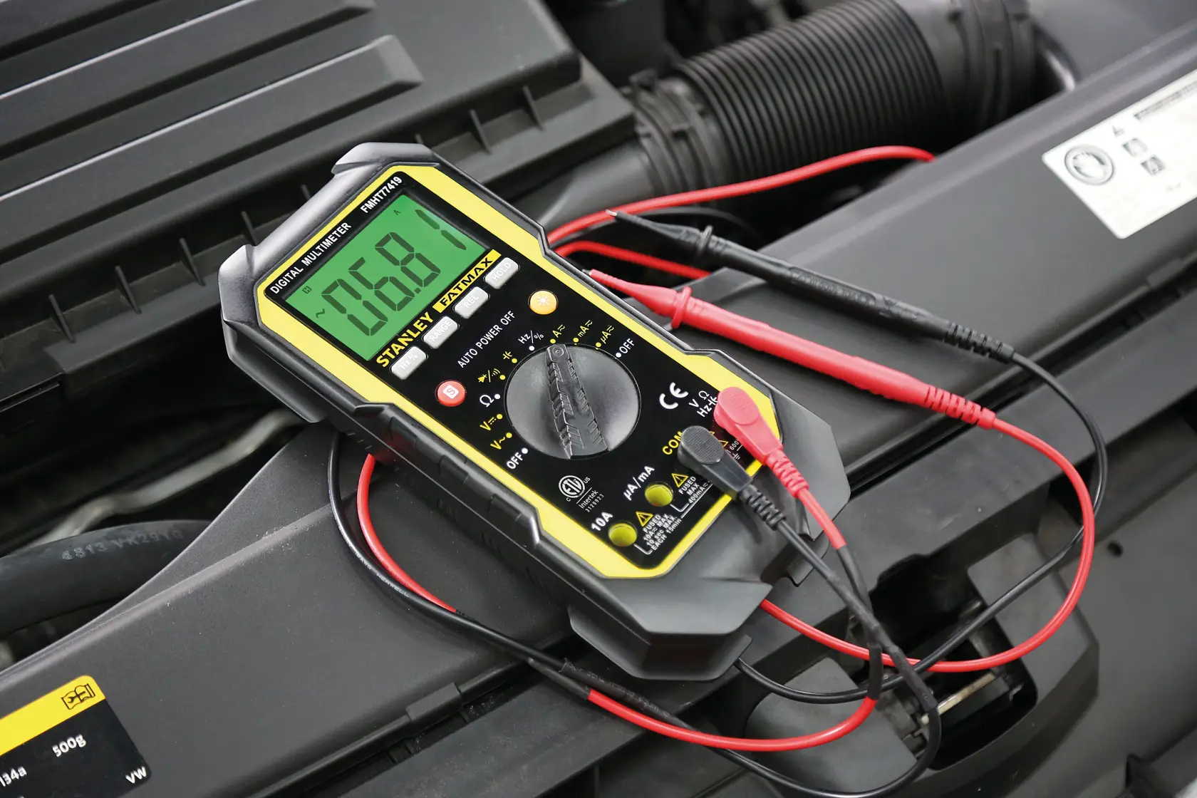 STANLEY FMHT0-77419 digital multimeter with yellow accents, shown on a car engine during electrical testing.