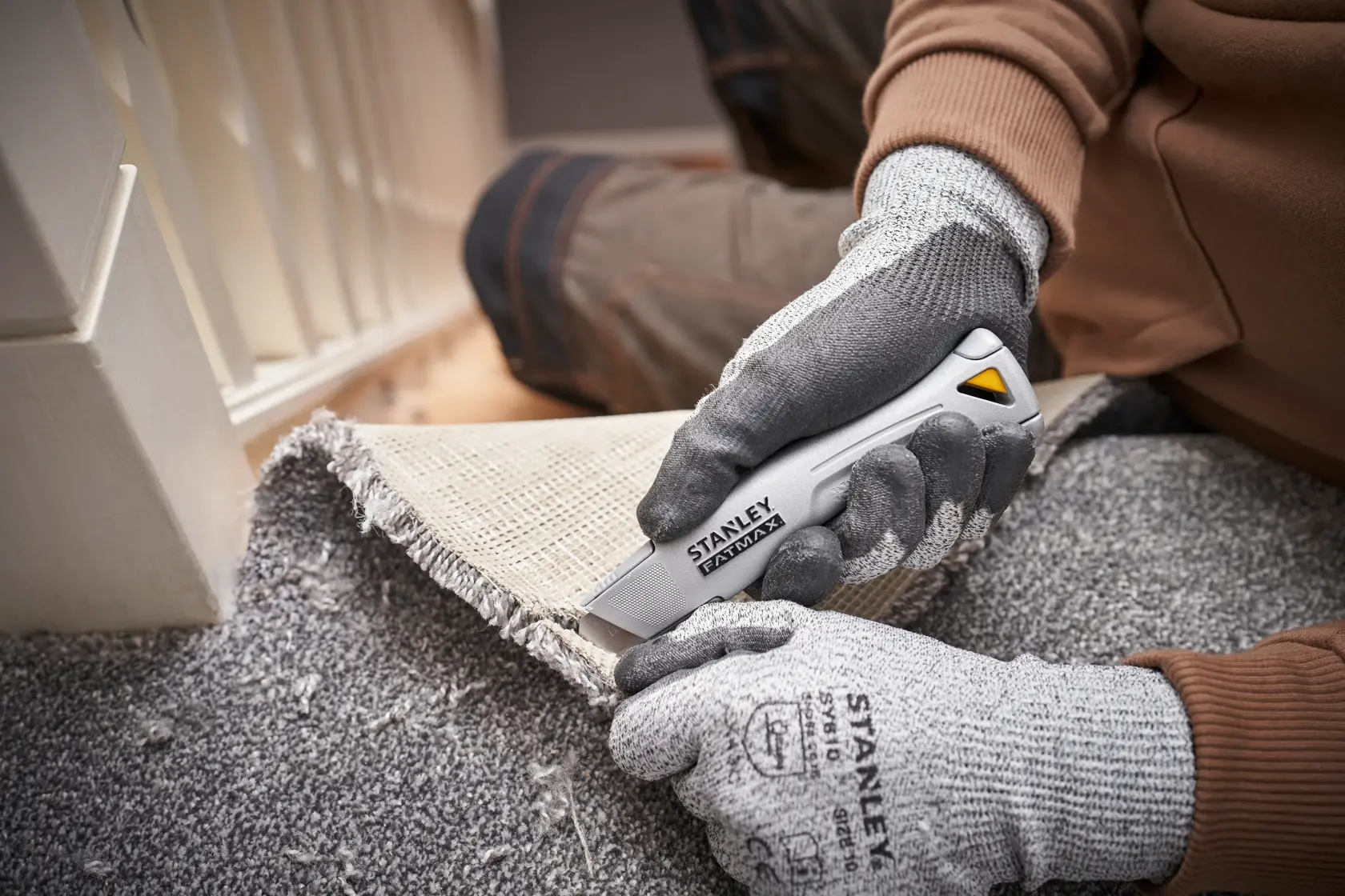 STANLEY FMHT10501-0 utility knife cutting carpet, photographed close-up in use with gloves.