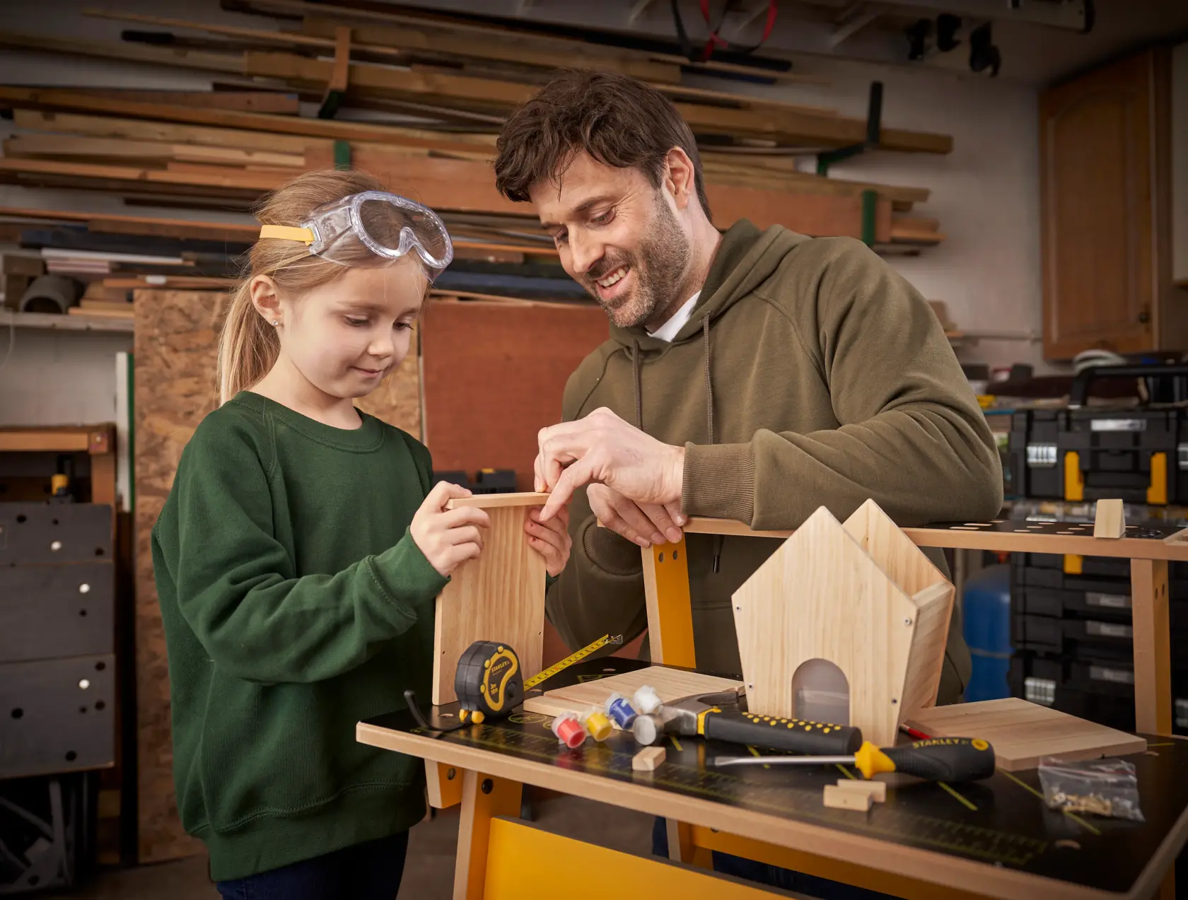 STANLEY K020-SY tape measure and screwdriver on workbench with wooden birdhouse and craft supplies, workshop setting.