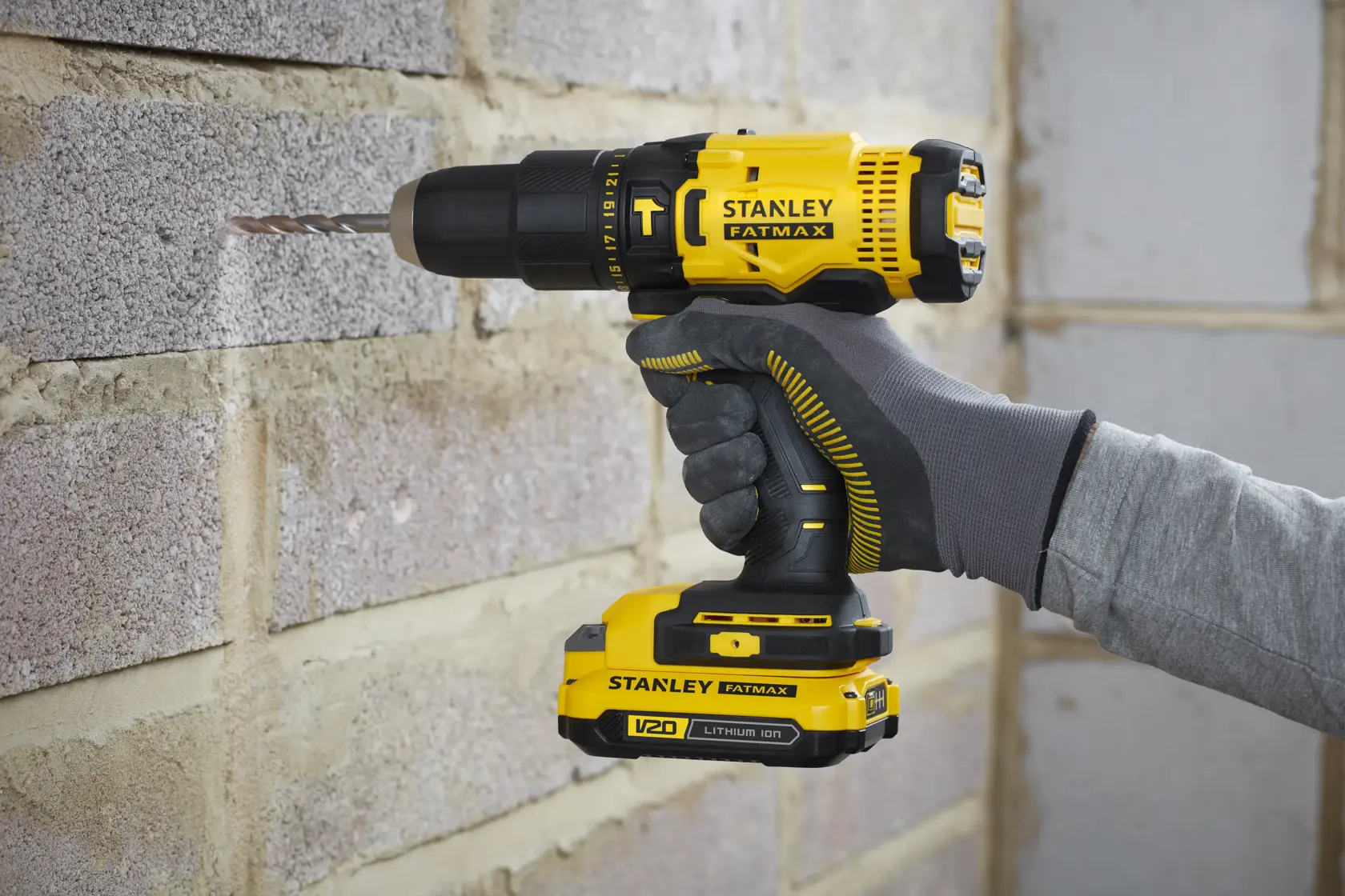 Stanley SFMCD711D21 cordless drill, yellow and black, in use drilling into brick wall, close-up side view.