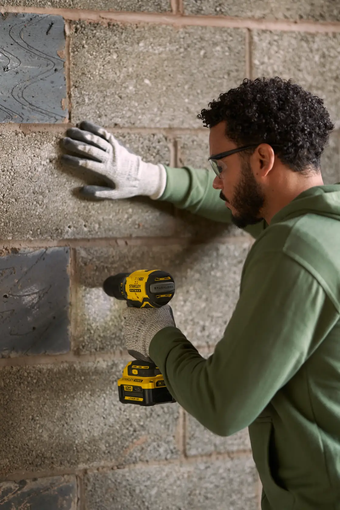 Photo of person using STANLEY SFMCK461M2S cordless drill on concrete wall, wearing gloves and green sweatshirt.