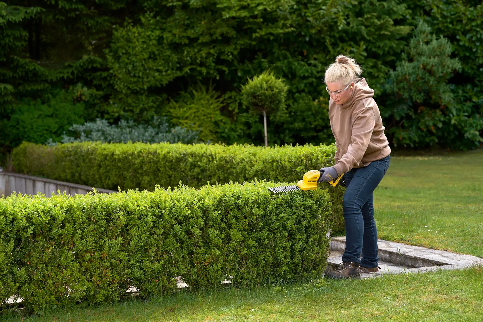 Photo of a person trimming a hedge with a STANLEY STCSS20D1 hedge trimmer in a garden.