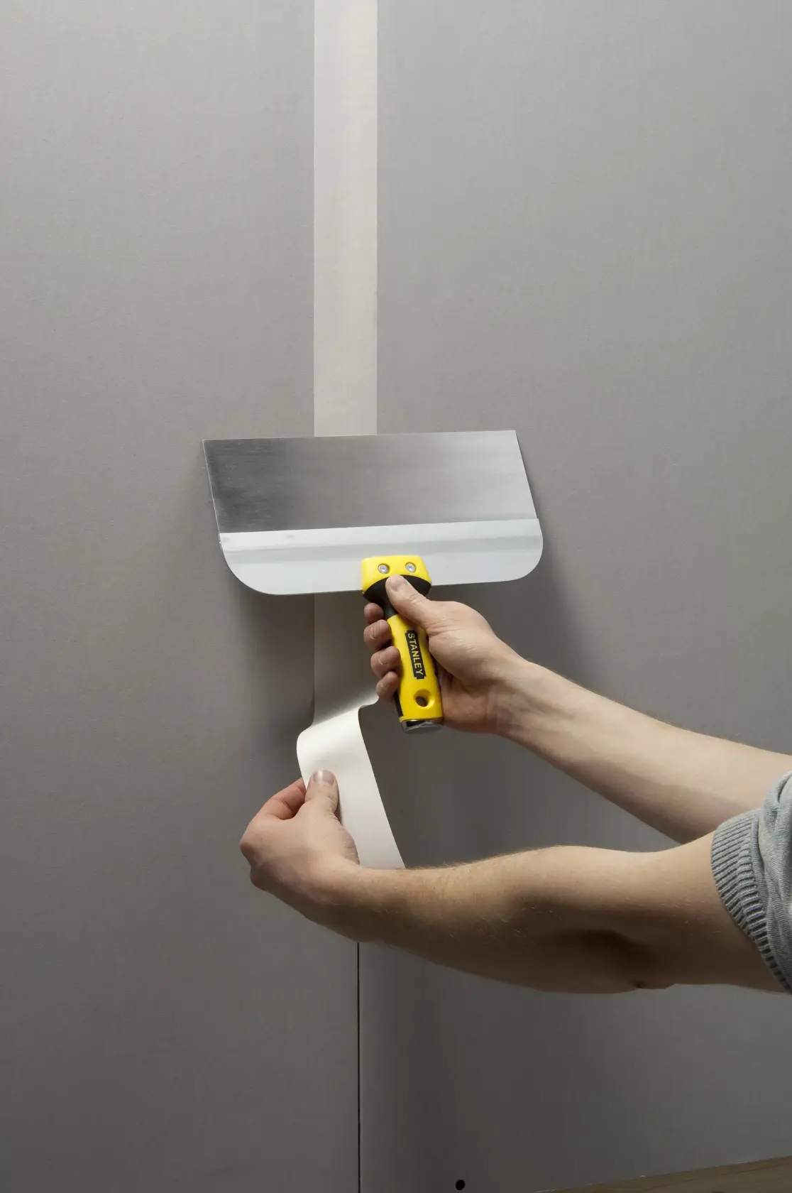 Photo of STANLEY STHT0-05771 drywall taping knife with yellow handle applying joint tape to plasterboard wall.